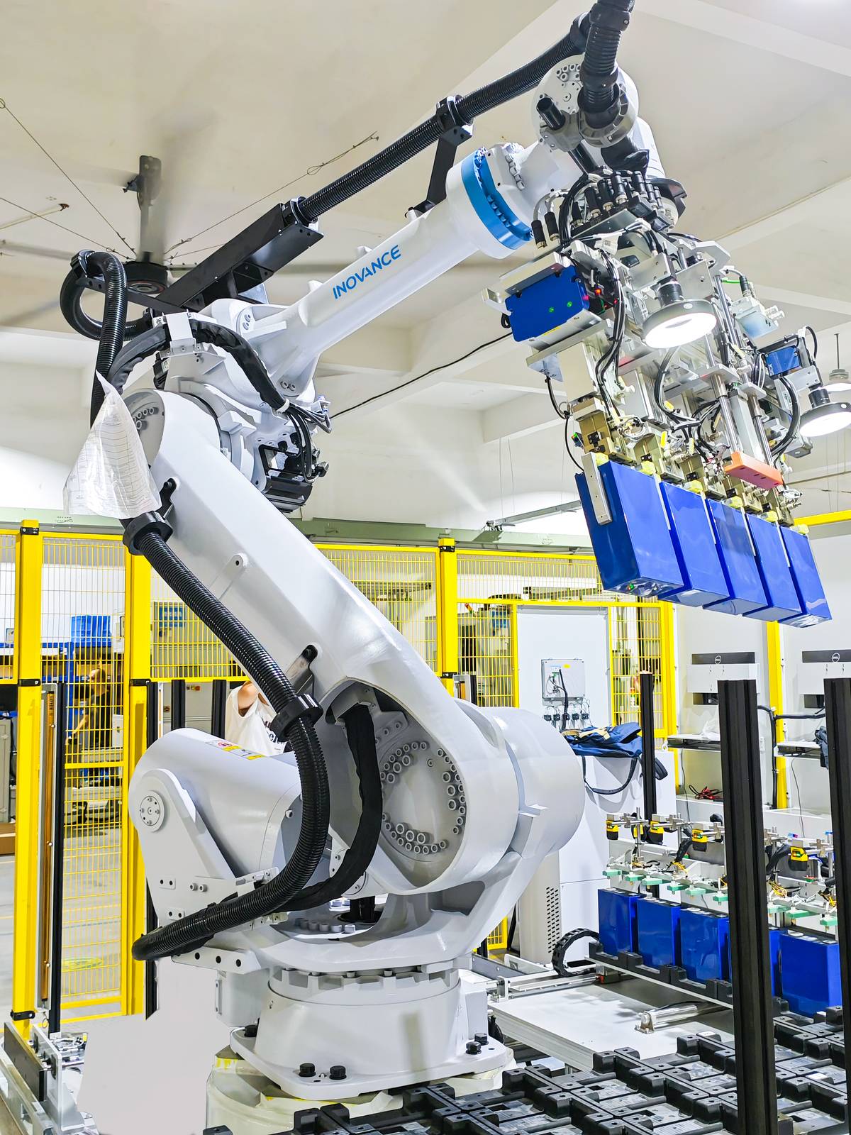 Robotic arm lifting blue battery modules over a tray of cells